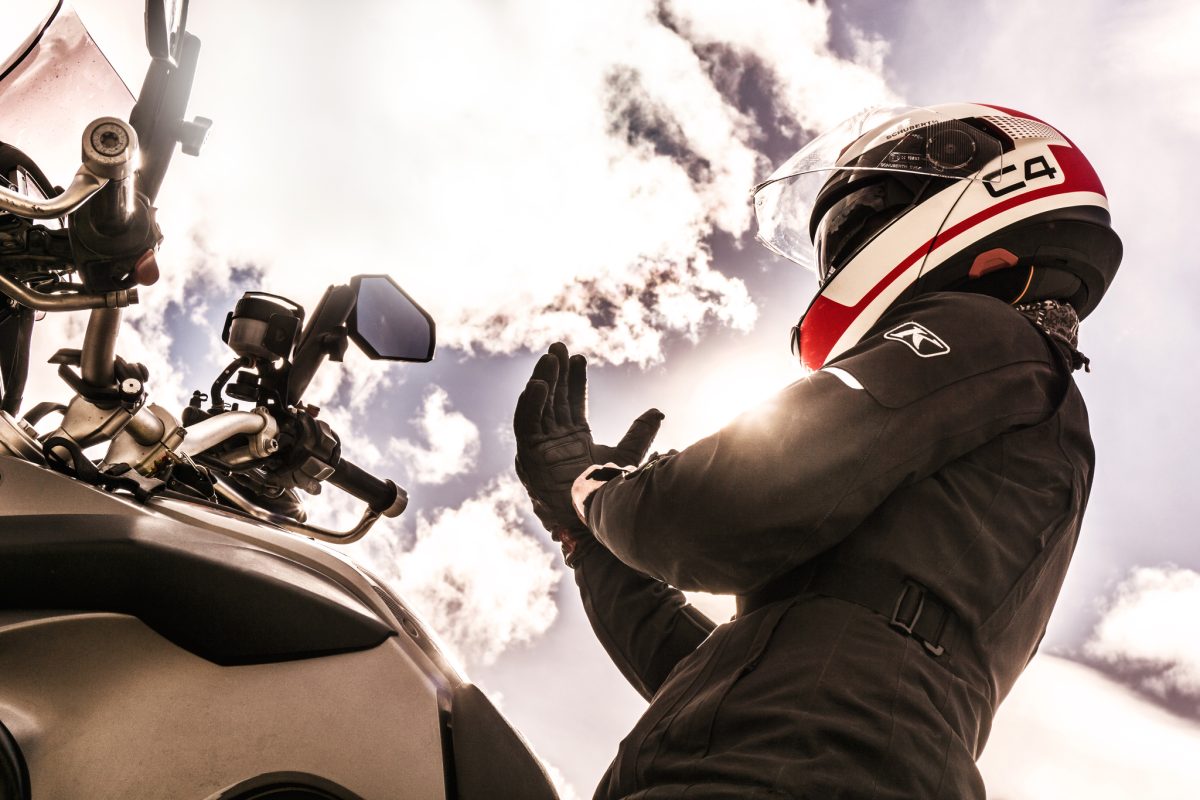 Motard levant les mains sur sa moto, casque intégral blanc rouge et noir avec écran, à contre-jour sous un ciel ensoleillé